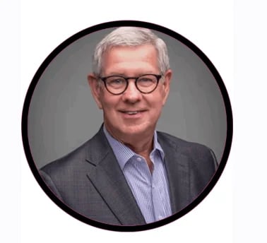 Professional headshot of a middle-aged man with gray hair and glasses wearing a blue striped shirt and gray blazer